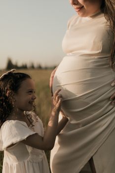 A young girl joyfully touching her pregnant mother's belly in a sunny outdoor setting. - Fotografer Sidoarjo