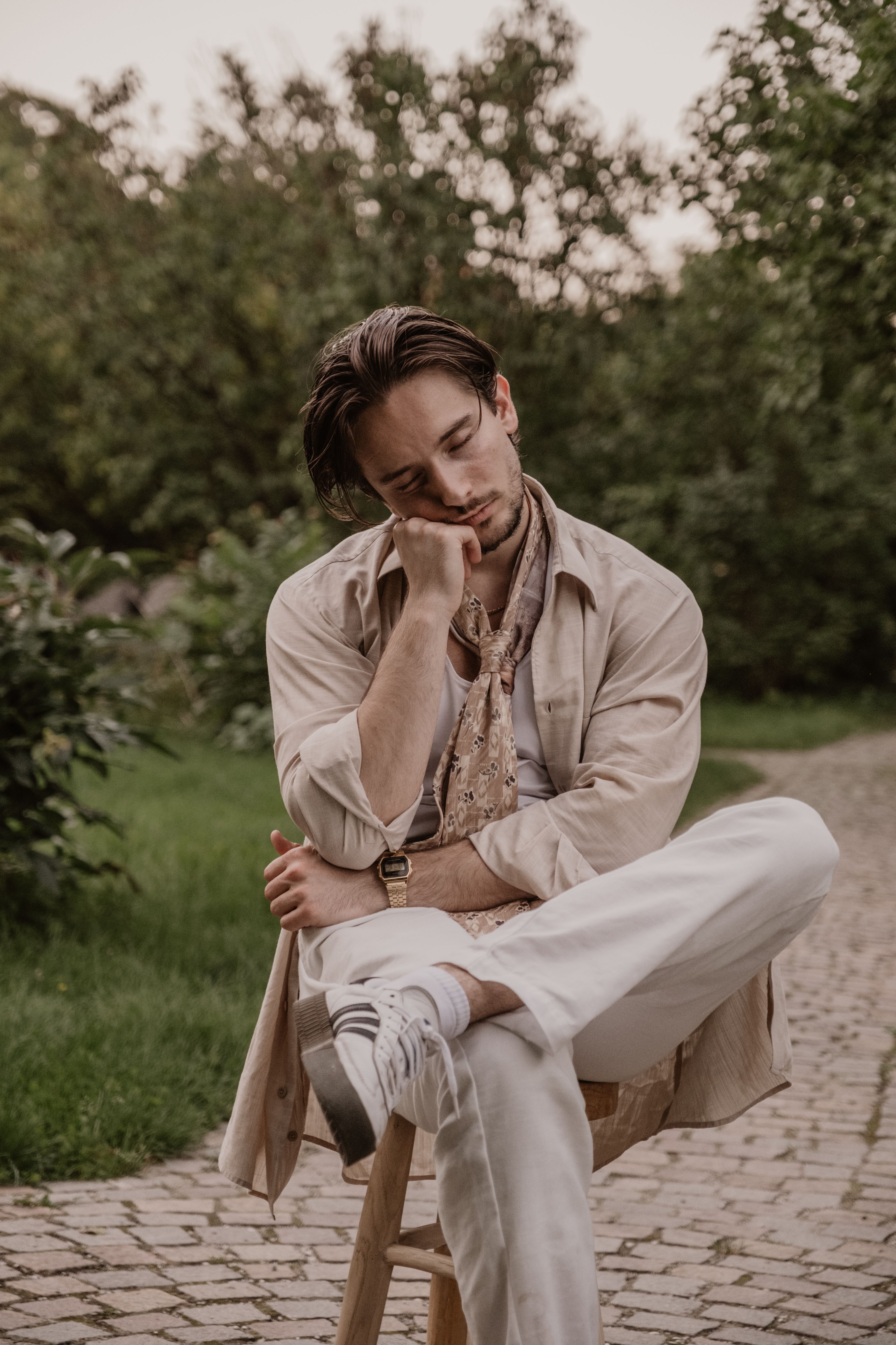 A fashionable young man sitting on a stool in a peaceful garden setting in Munich during the day.