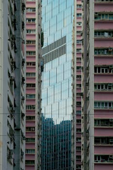 Modern glass skyscraper flanked by urban buildings, showcasing reflection and geometry.