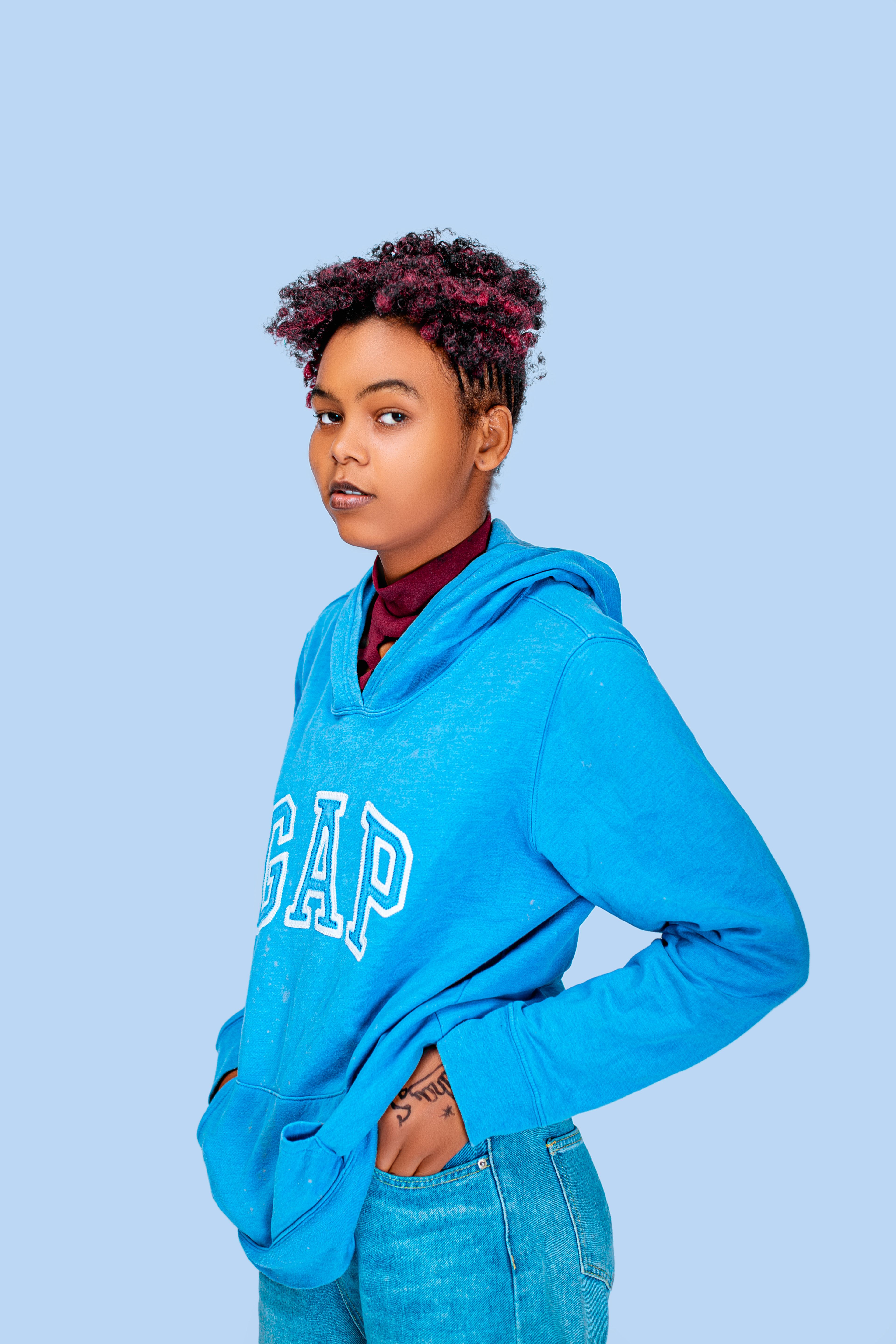 Fashion portrait of a confident young woman in a blue hoodie standing against a pastel background.