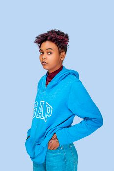 Fashion portrait of a confident young woman in a blue hoodie standing against a pastel background.