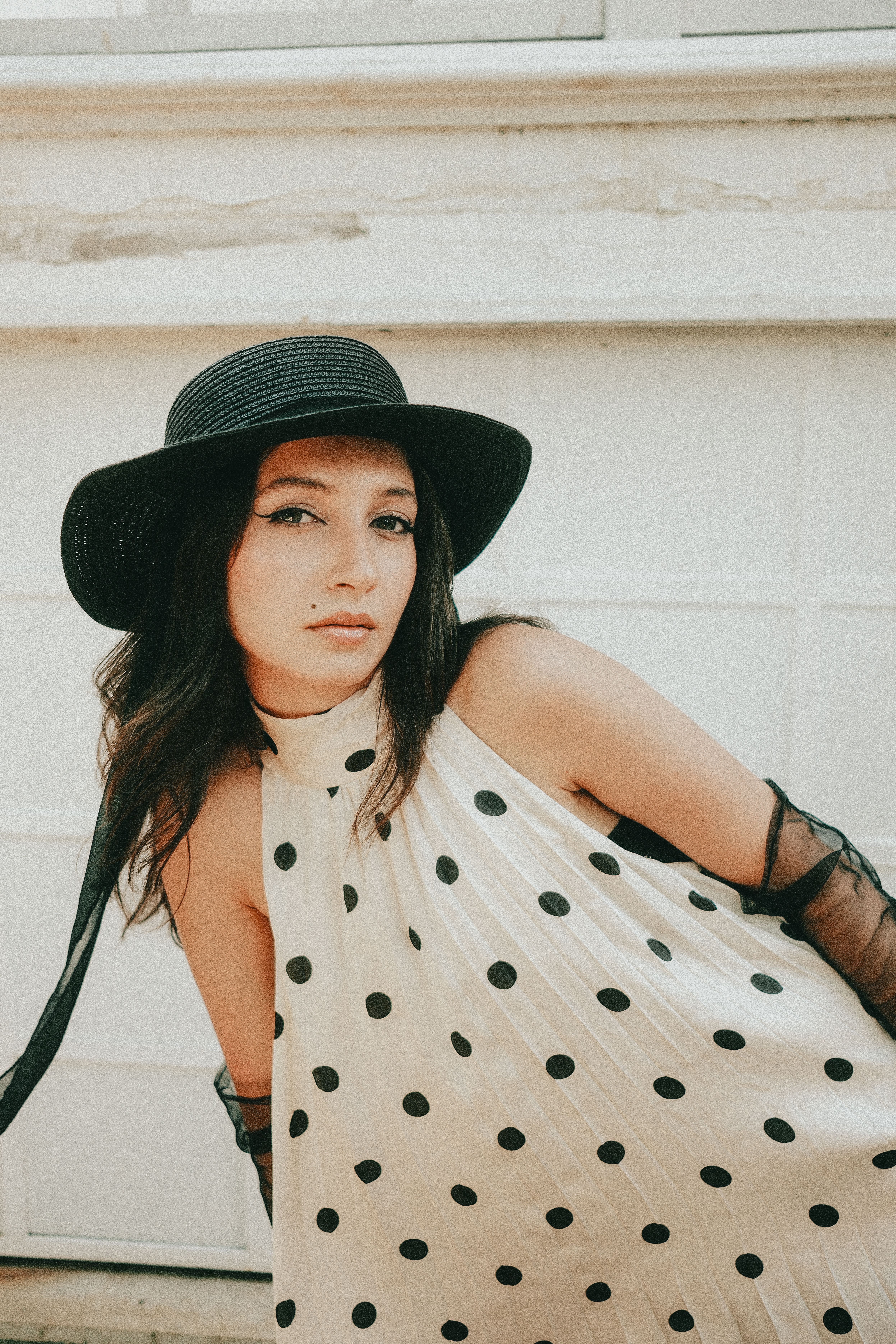 Elegant woman wearing polka dot dress and black hat poses outdoors.