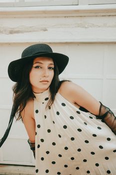 Elegant woman wearing polka dot dress and black hat poses outdoors.
