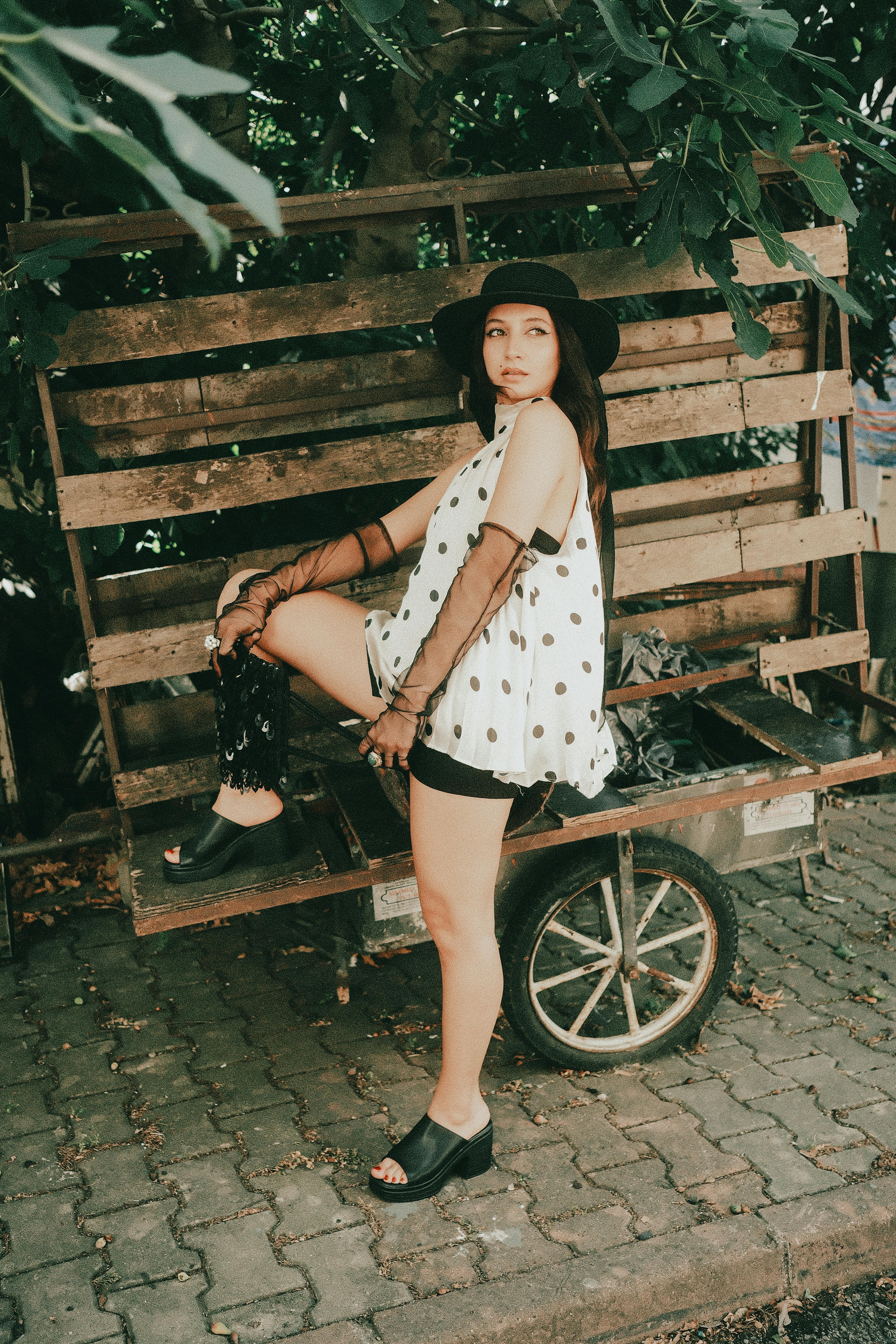 Fashionable woman in polka dot top and hat posing on rustic cart outdoors.