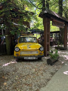 A vintage yellow car parked at the entrance of a rustic outdoor pizzeria surrounded by greenery.