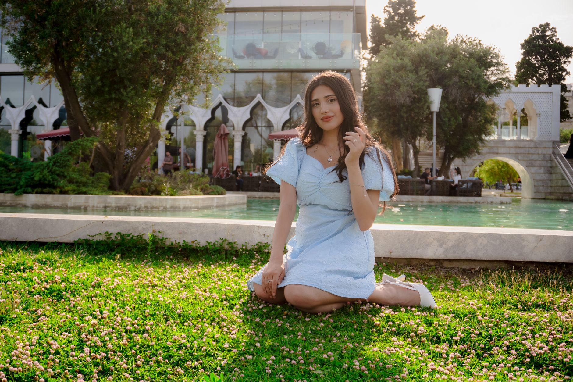 Elegant young woman in a blue dress poses in a Baku park near a fountain, exuding beauty and style.