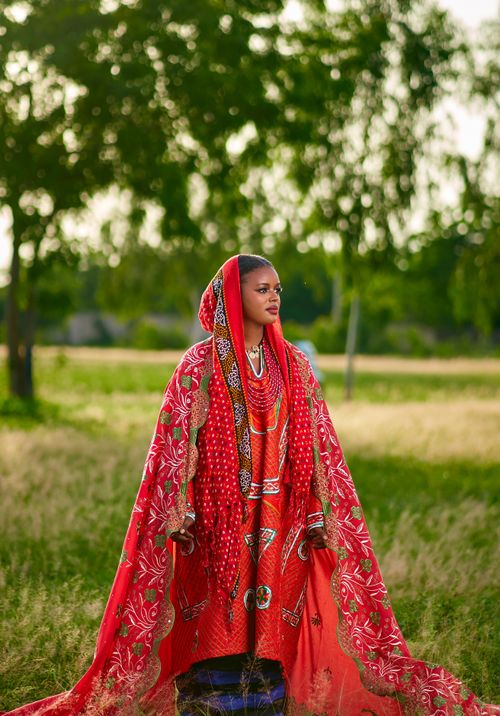 Free A woman in vibrant red traditional attire stands elegantly in a lush outdoor setting. Stock Photo