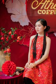 Stylish portrait of a young Asian woman in a traditional floral red dress with braided hair, seated in a festive setting.