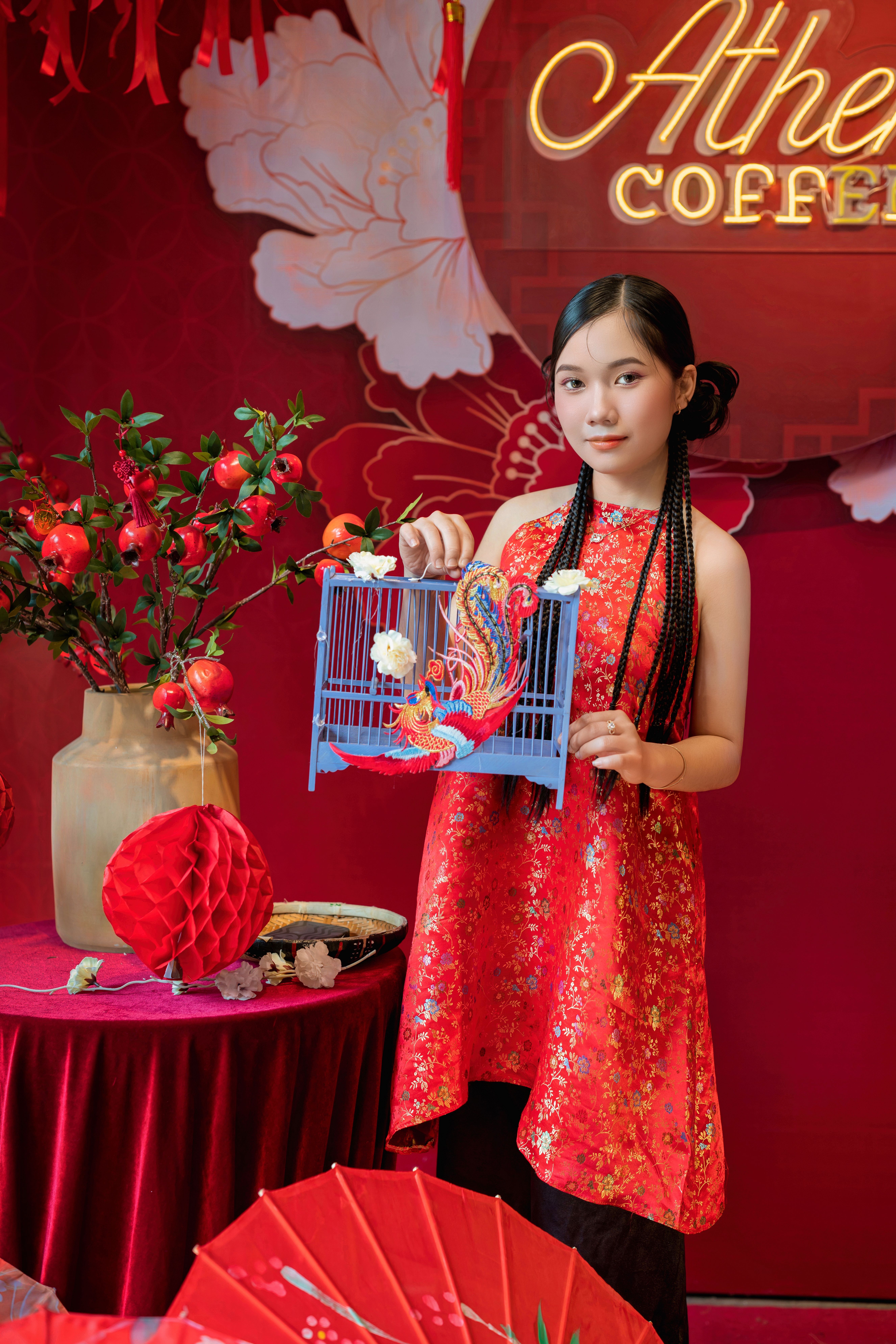 Girl in traditional attire holding a birdcage, surrounded by red festive decorations.