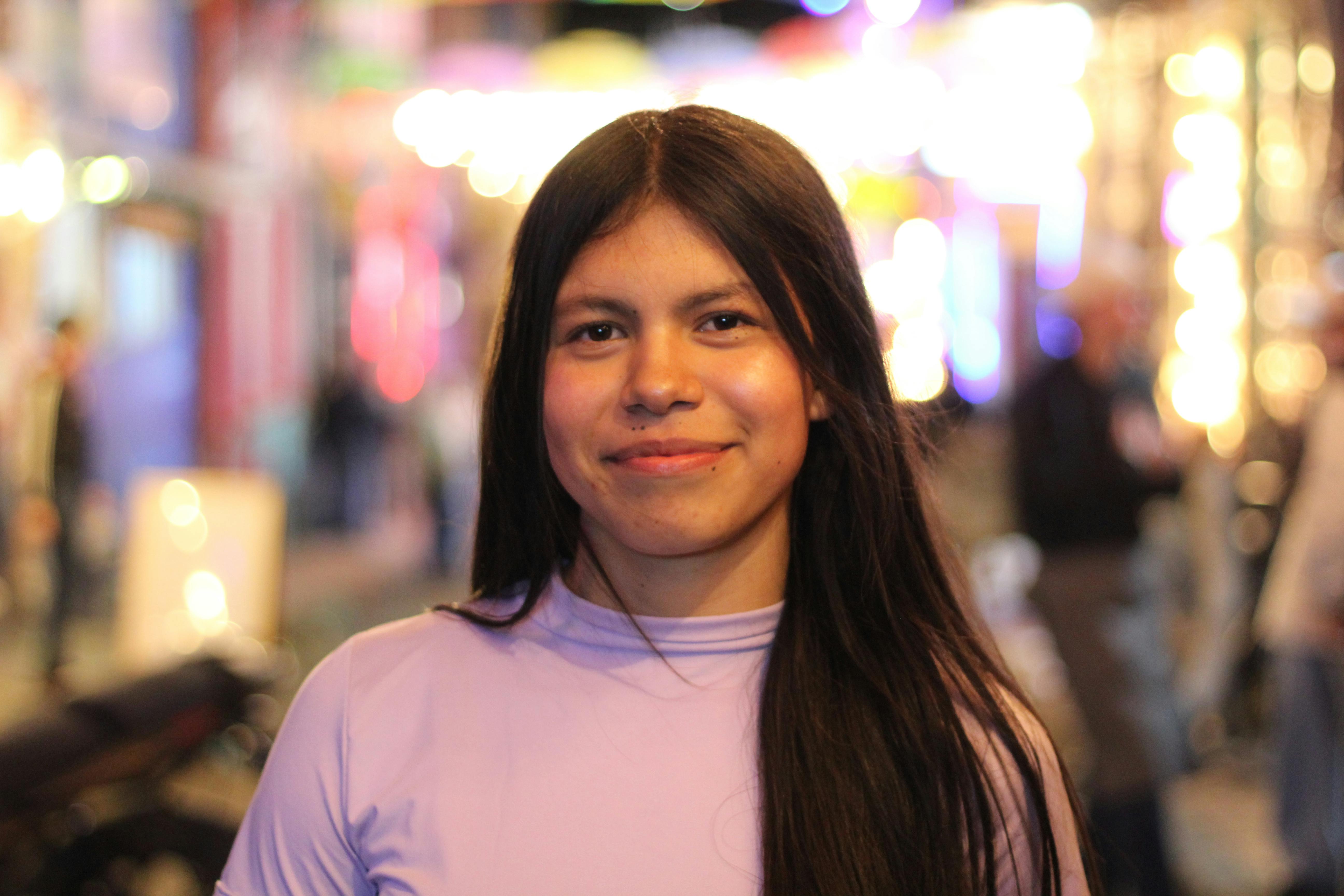 Portrait of a young woman smiling outdoors at night with lively street lights in the background.