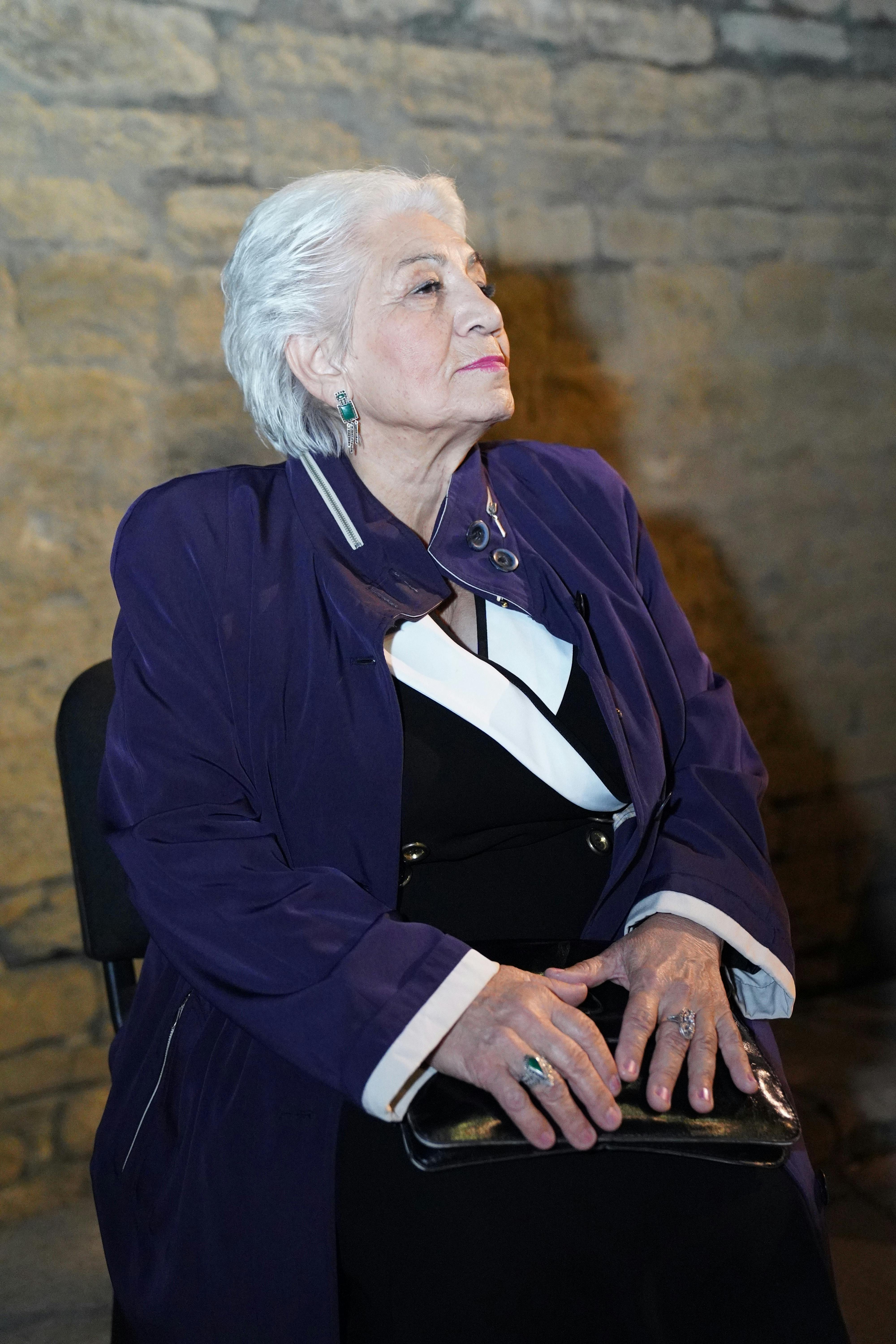 Elderly woman dressed elegantly in purple and white sits indoors against a stone wall.