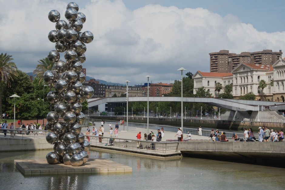 Una escultura moderna junto al río en Bilbao, España, que presenta esferas brillantes con una ciudad de fondo.