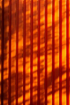 Vertical lines with warm orange hues create abstract light and shadow patterns on a textured surface.