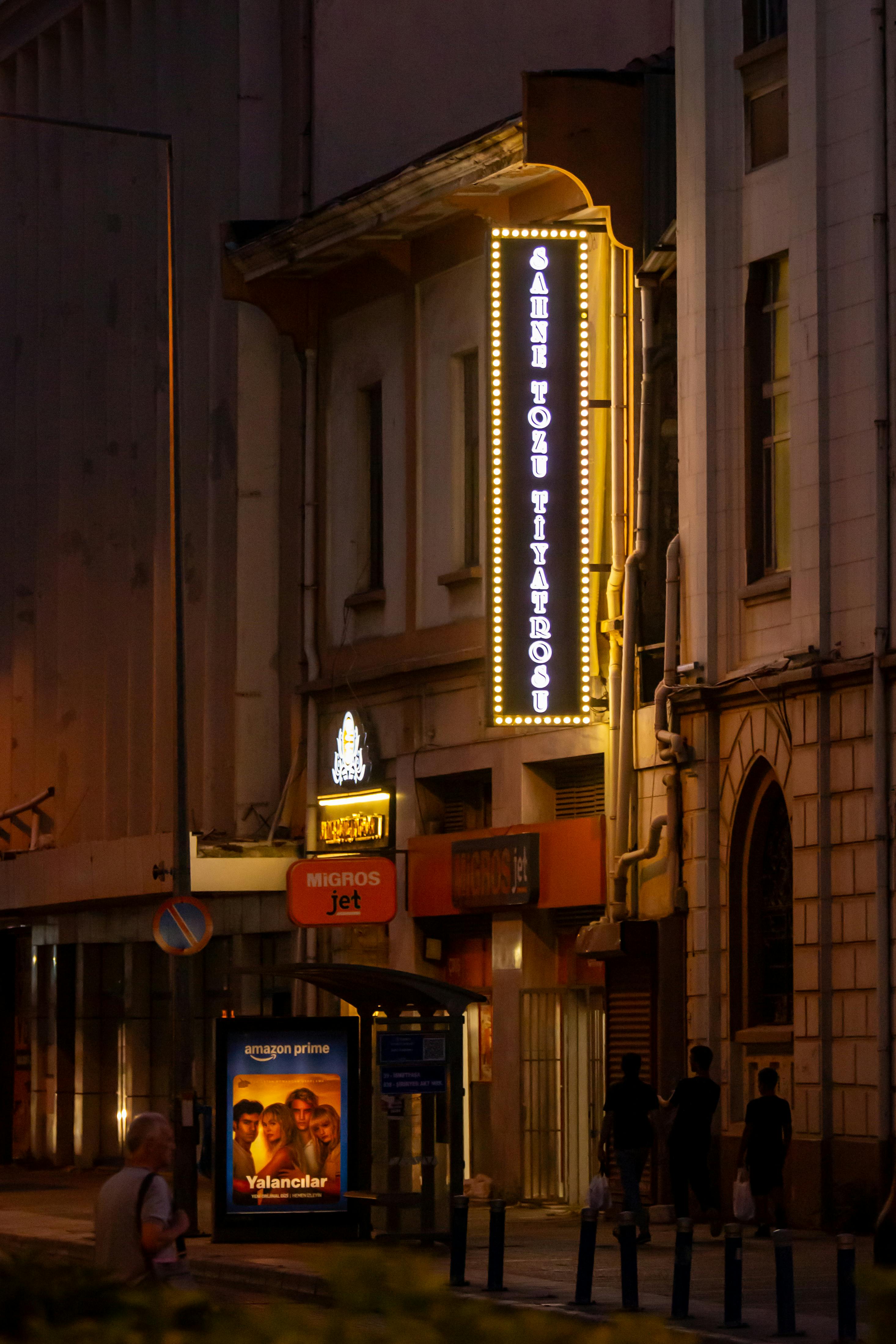 Bezpłatne Urokliwy nocny widok ulicy w Izmirze. Widać podświetlone szyldy teatru i spacerujących ludzi. Zdjęcie z galerii