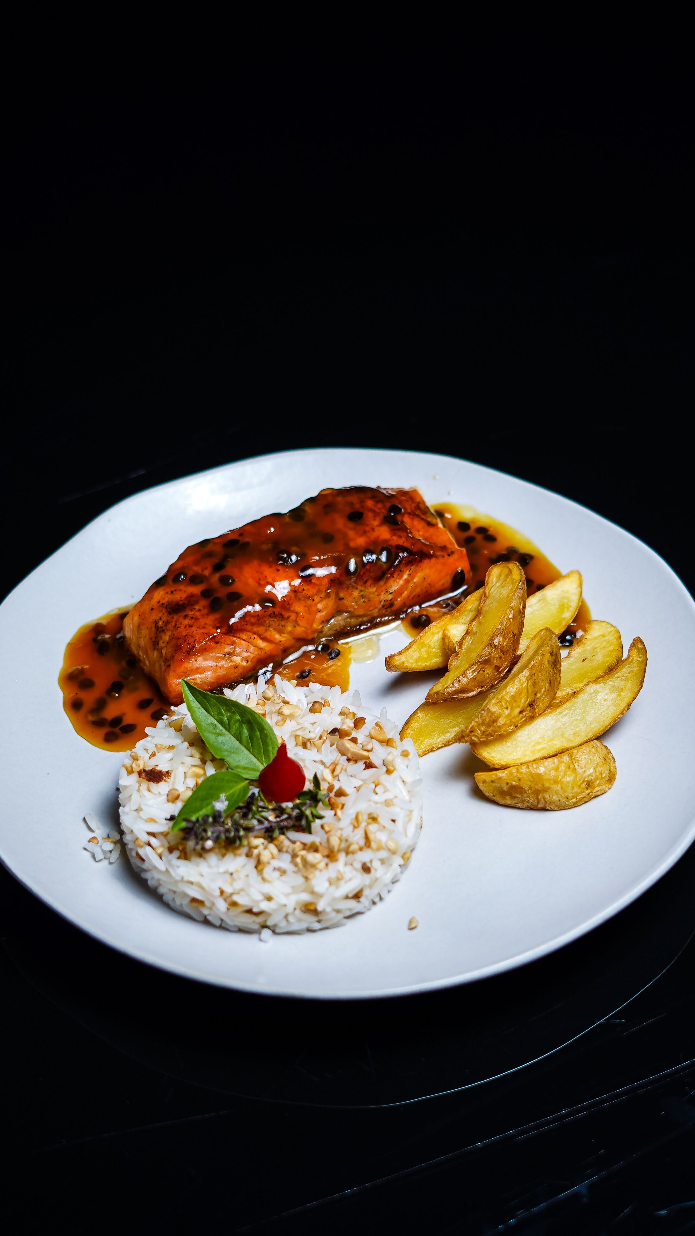 Delicious plated salmon with sauce, rice, and potatoes on a black background.