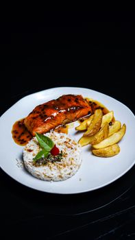 Delicious plated salmon with sauce, rice, and potatoes on a black background.