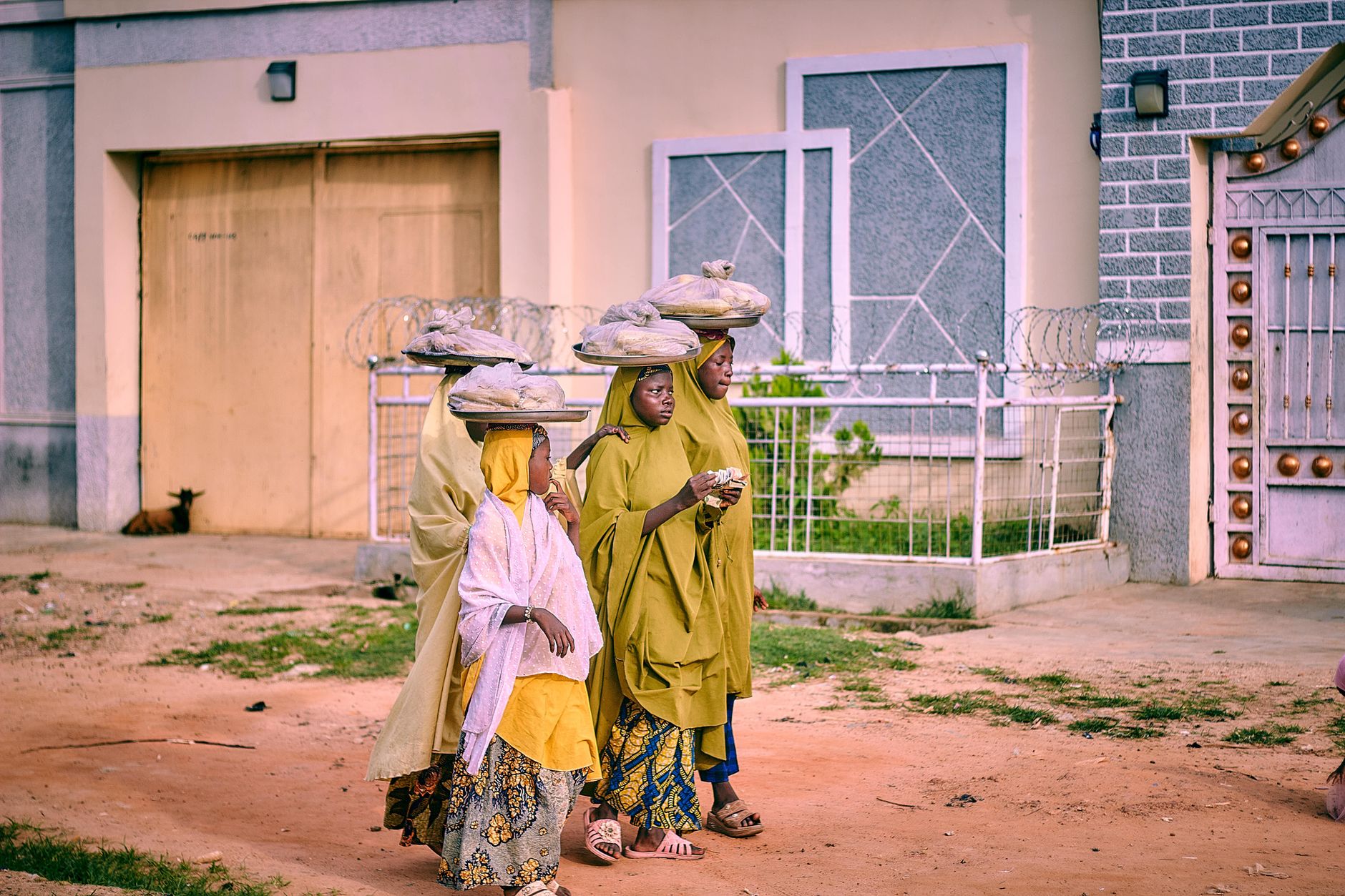 Three women in colorful traditional African attire walk outdoors, carrying items on their heads.