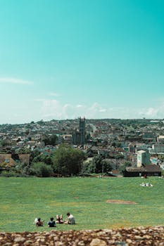 A picturesque view of Cork cityscape with people relaxing on a grassy hill under a clear blue sky.