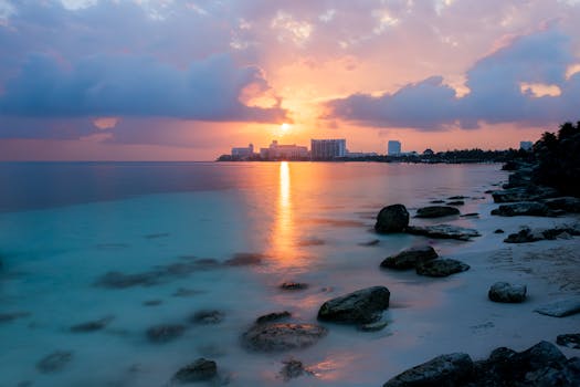 Serene sunrise over Cancun's rocky beach with a view of the Caribbean Sea and distant hotels.