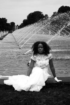 A woman in a white dress poses by a fountain, captured in black and white photography.