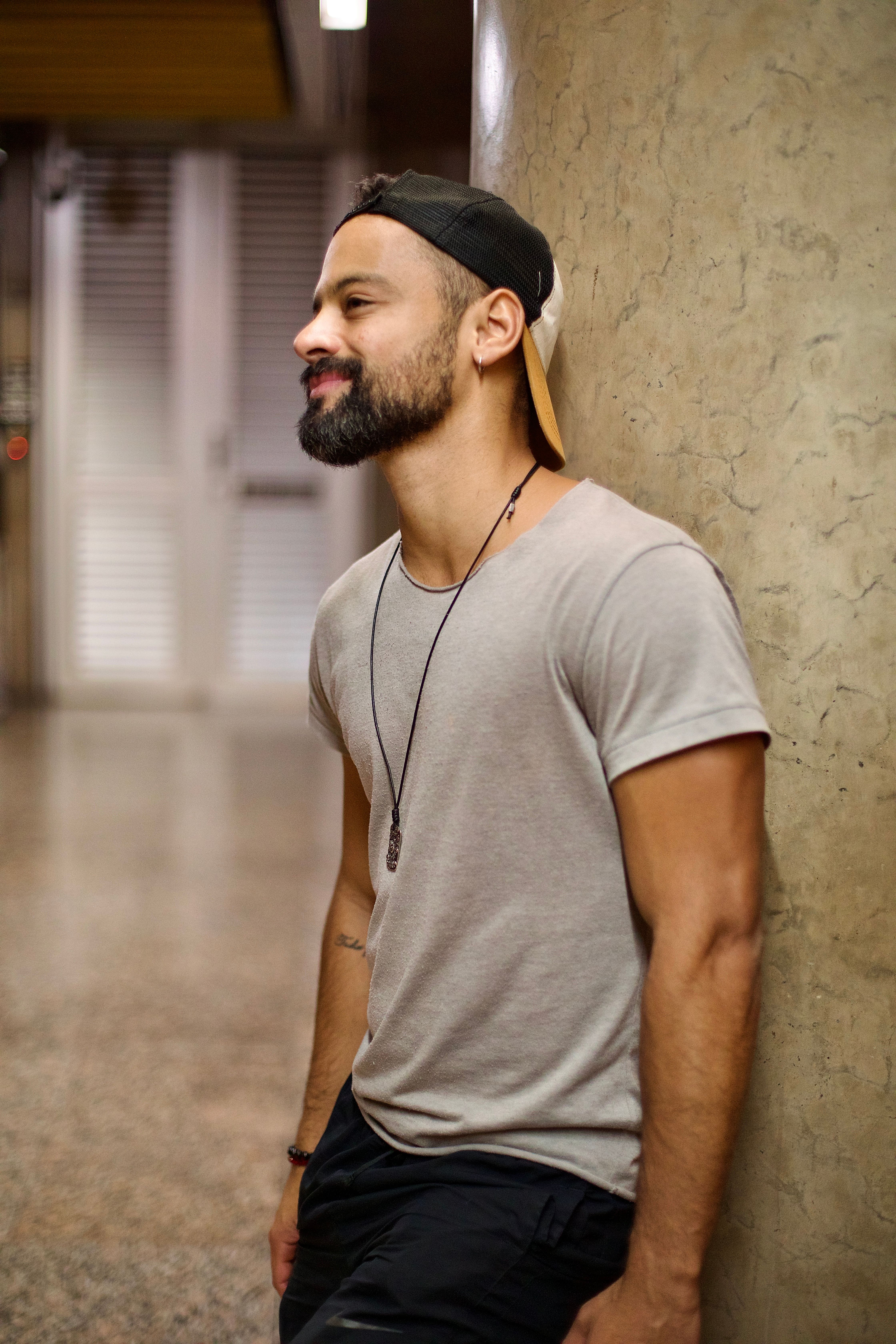 A young man leaning against a wall, wearing a cap and casual attire, indoors.