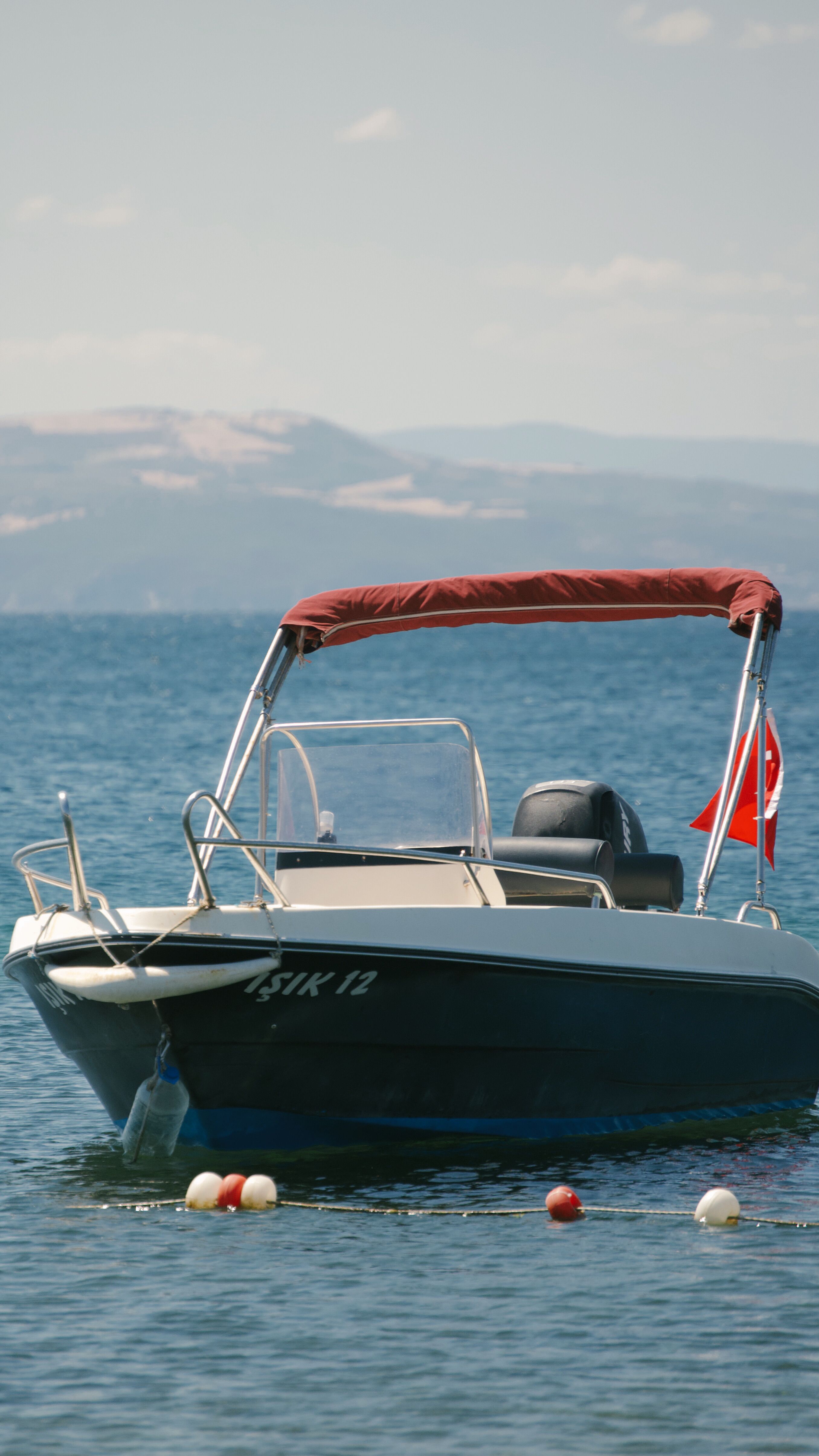 A small boat with a Turkish flag floats on a clear blue sea, mountains in the background.