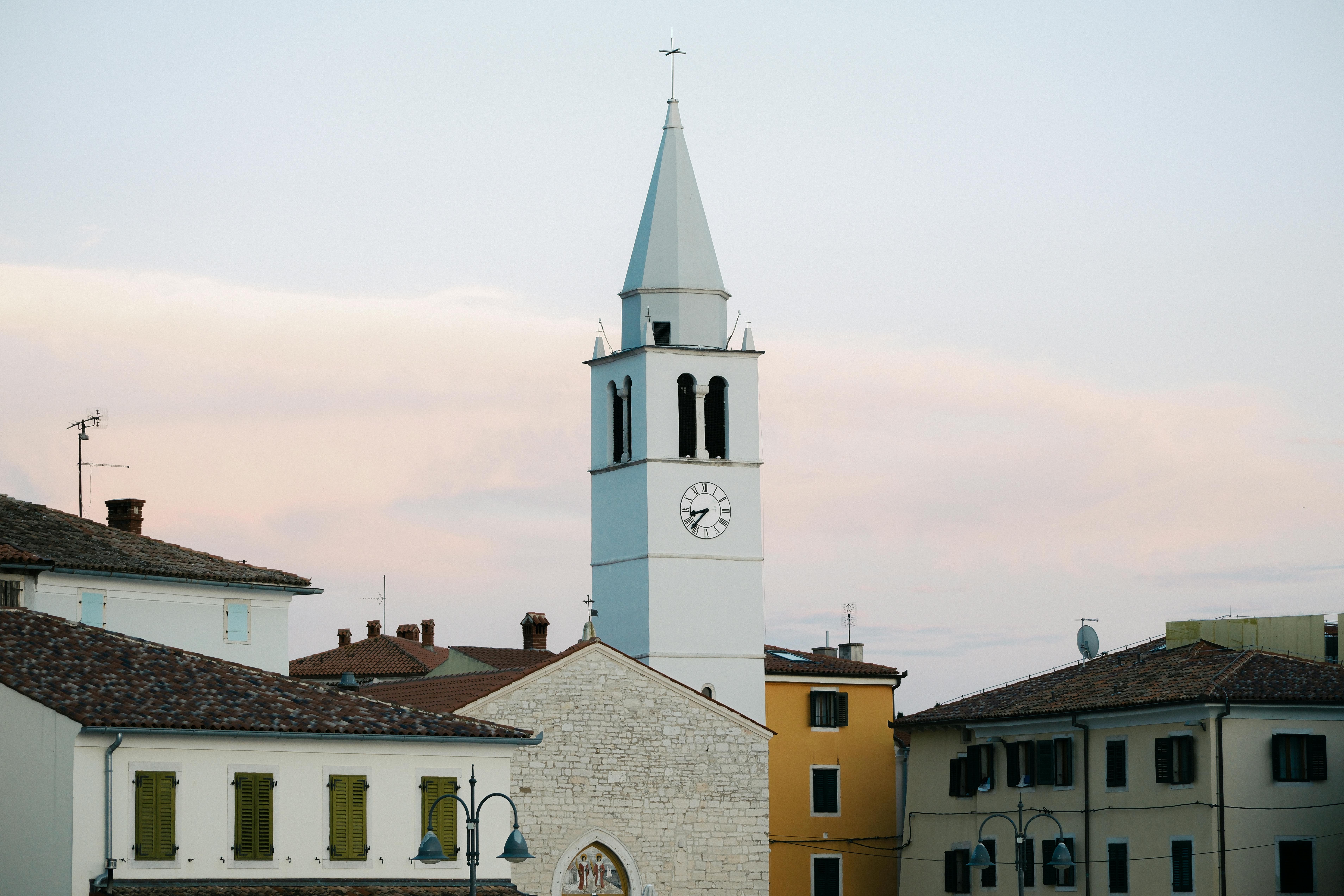 Beautiful Croatian village church steeple with clock against the evening sky, framed by historic buildings.