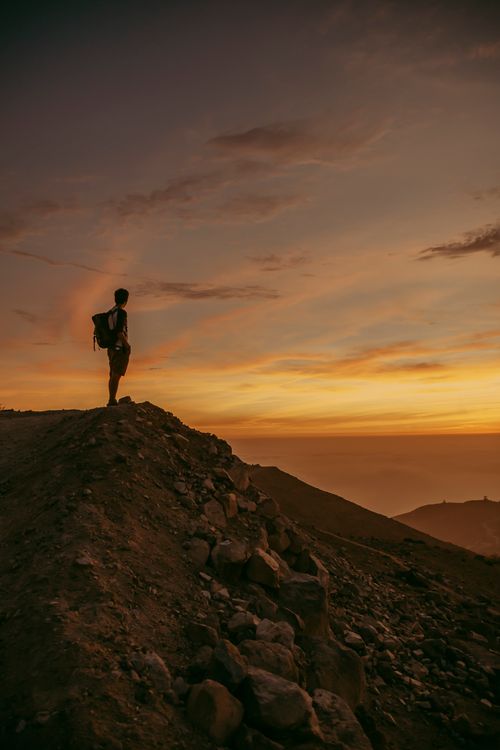 Free A lone hiker stands atop a rocky mountain, silhouetted against a dramatic sunset sky. Stock Photo