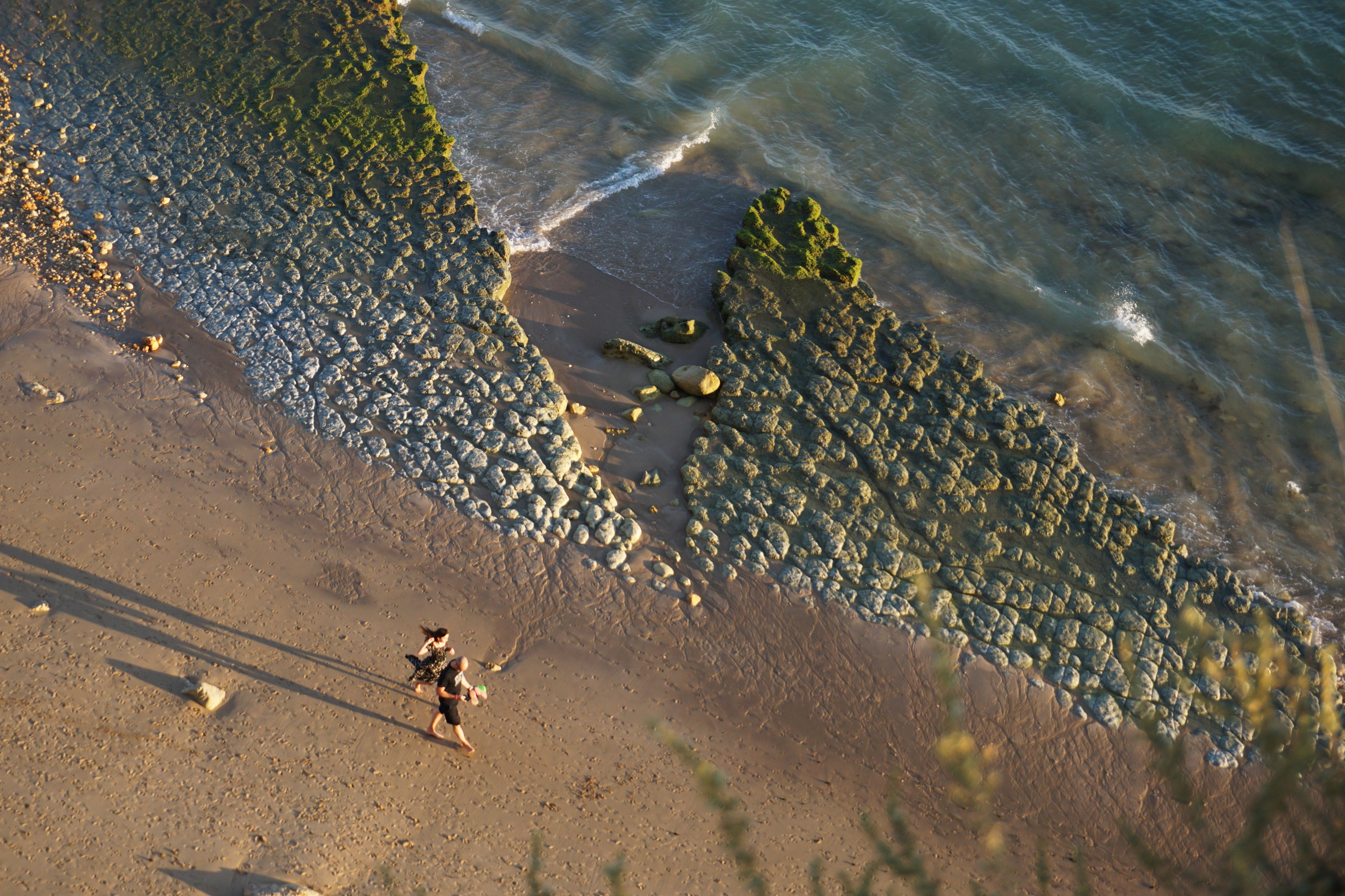 Aerial view of a beach with two people walking along coastal rocks and waves.