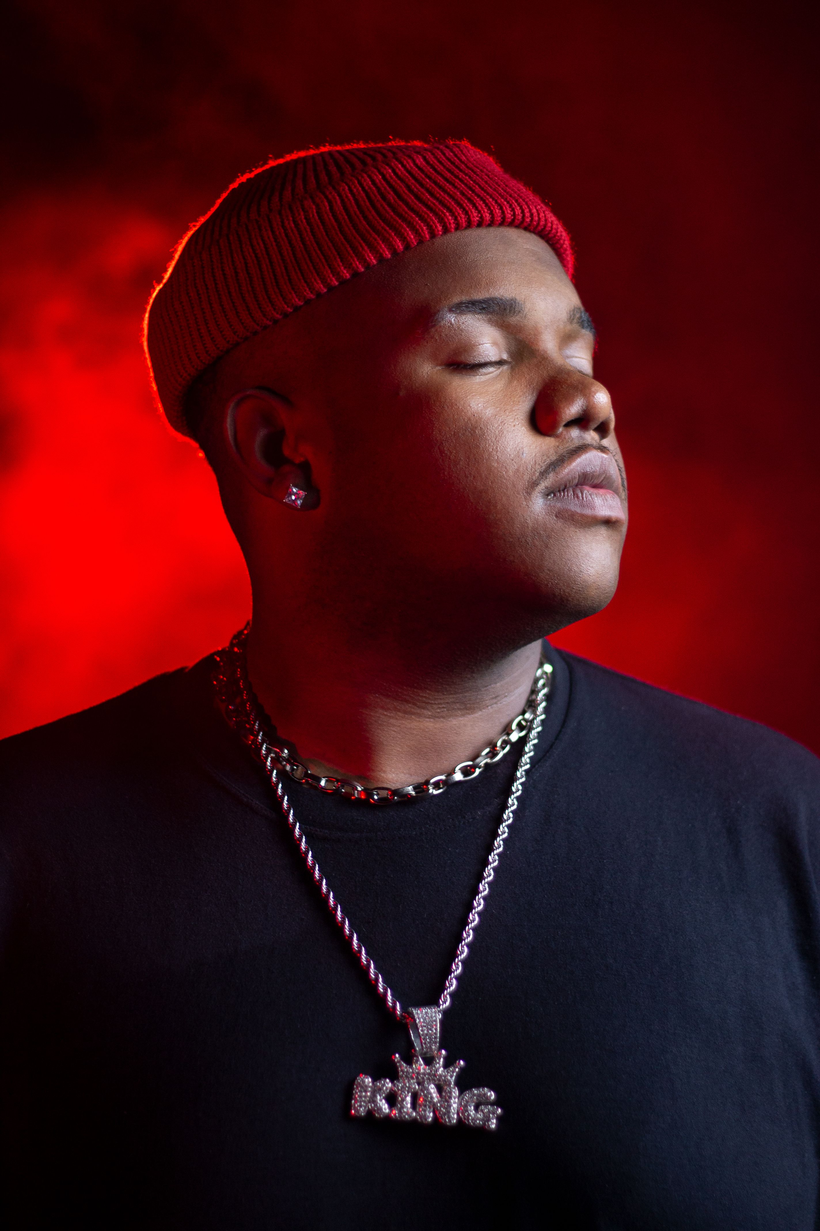 Confident man with closed eyes in a red beanie and black shirt, set against a vibrant red backdrop.