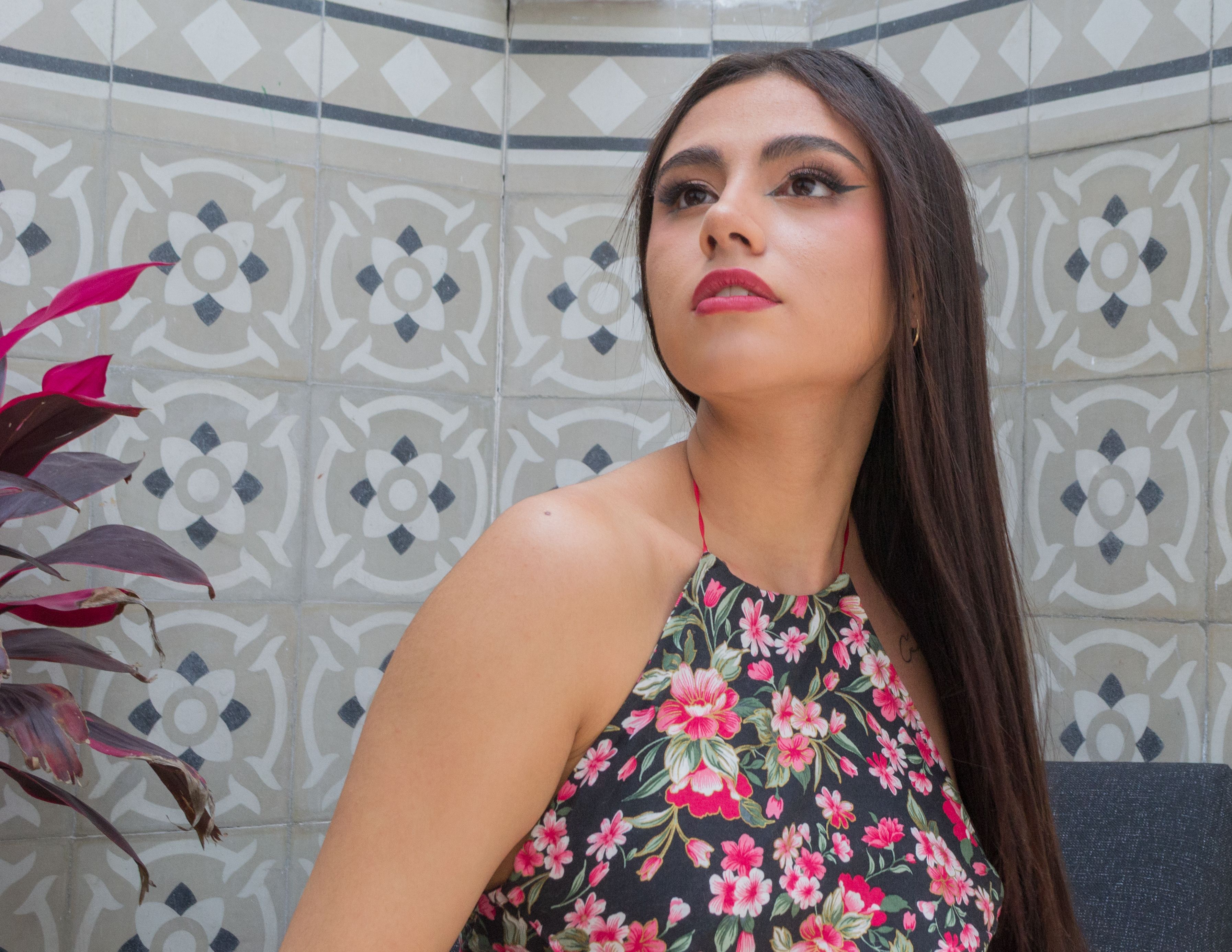 Mulher jovem com cabelos longos e escuros vestindo um top floral olhando para cima, contra um fundo de azulejos.