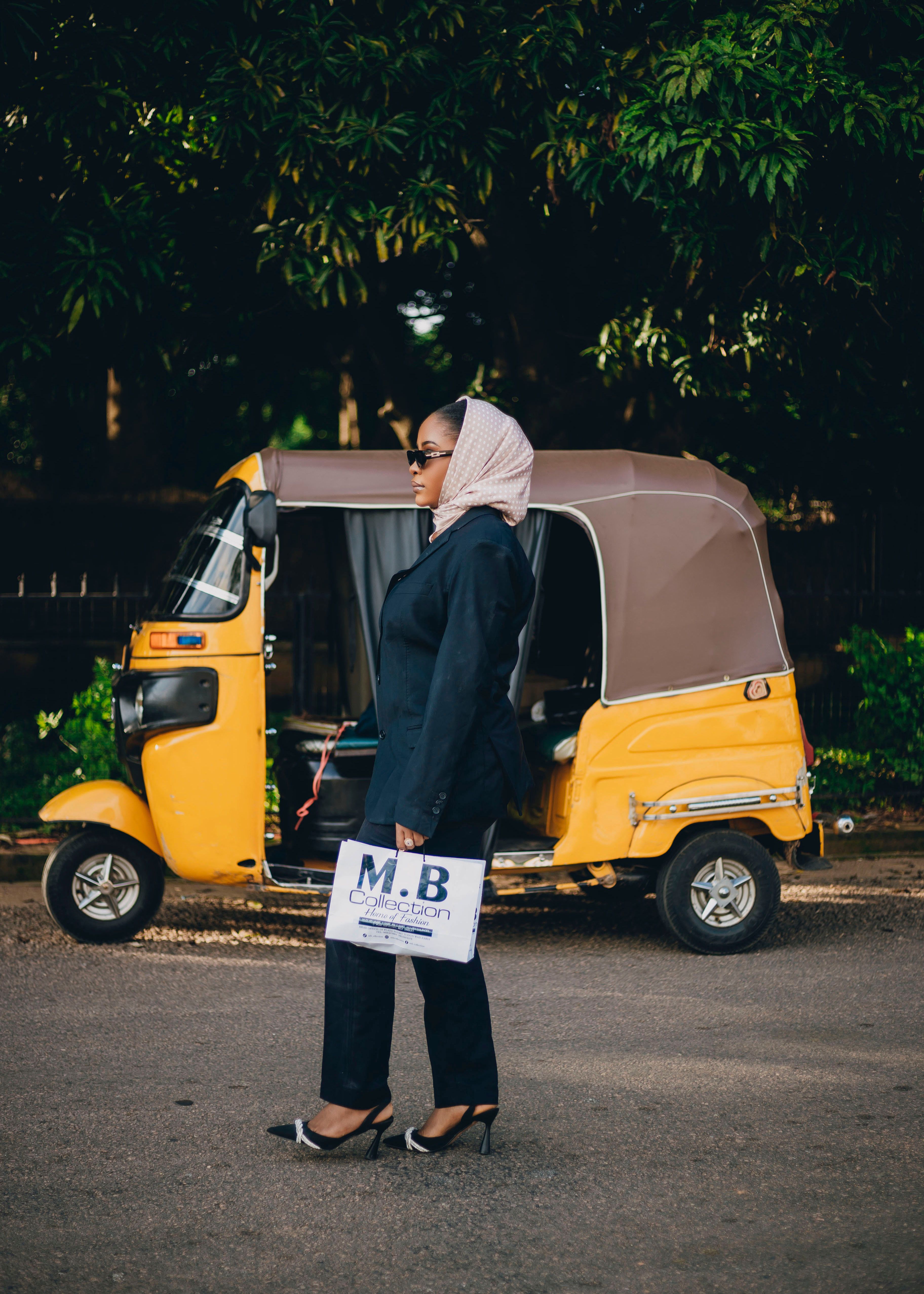 Fashionable woman with hijab and bag stands beside vibrant yellow auto rickshaw.