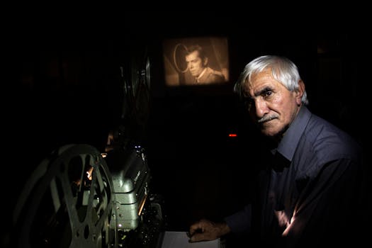 An elderly man stands next to a vintage film projector, casting a film in a dark room.