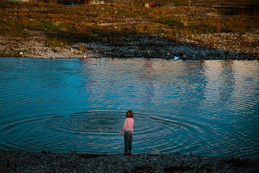 A young child in a striped shirt plays by the lakeside, rippling the calm water under a clear sky.