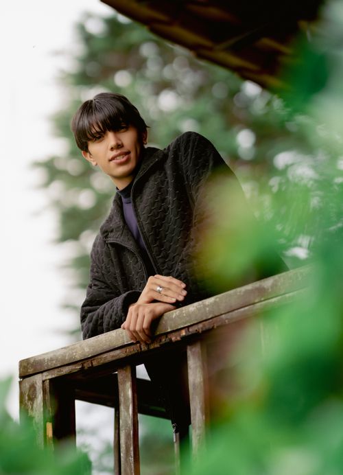 Free Young man in a dark jacket leaning on a wooden balcony railing surrounded by greenery. Stock Photo