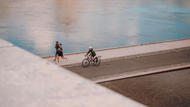 Two people running and a cyclist on a riverside path in an urban setting during the day.
