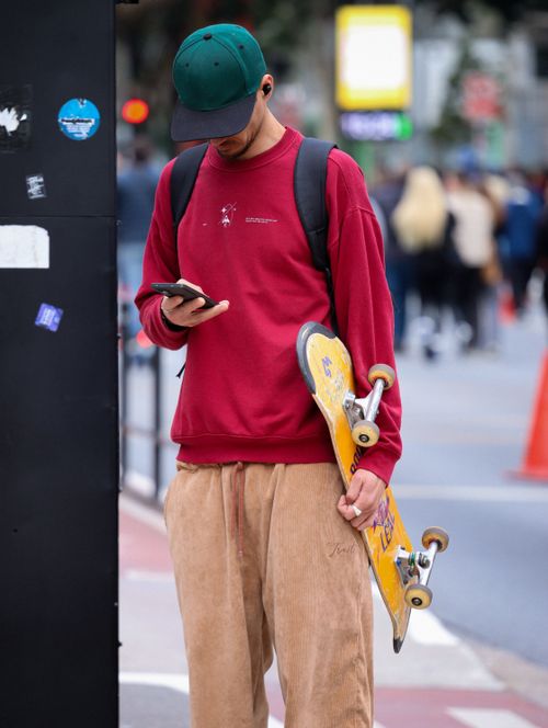 Free A young man holding a skateboard checks his phone on a busy city street, blending street fashion with urban culture. Stock Photo
