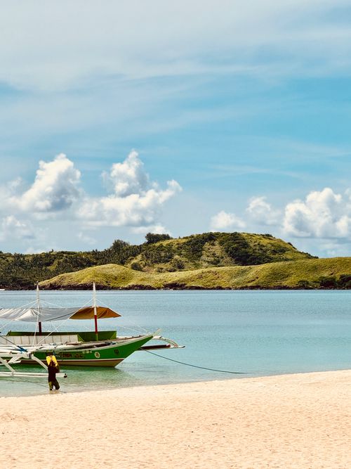 Free A serene tropical beach with a green banca boat and calm blue waters under a clear sky. Stock Photo