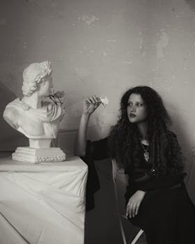 A woman holding a white rose beside a classical bust in a moody setting.