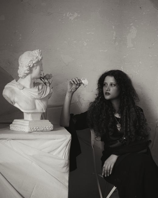 A woman holding a white rose beside a classical bust in a moody setting.