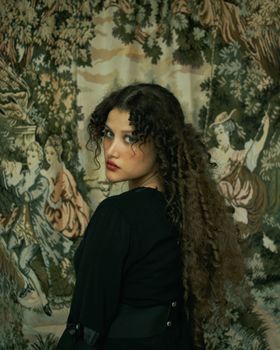 Elegant portrait of a woman with curly hair standing in front of a tapestry.