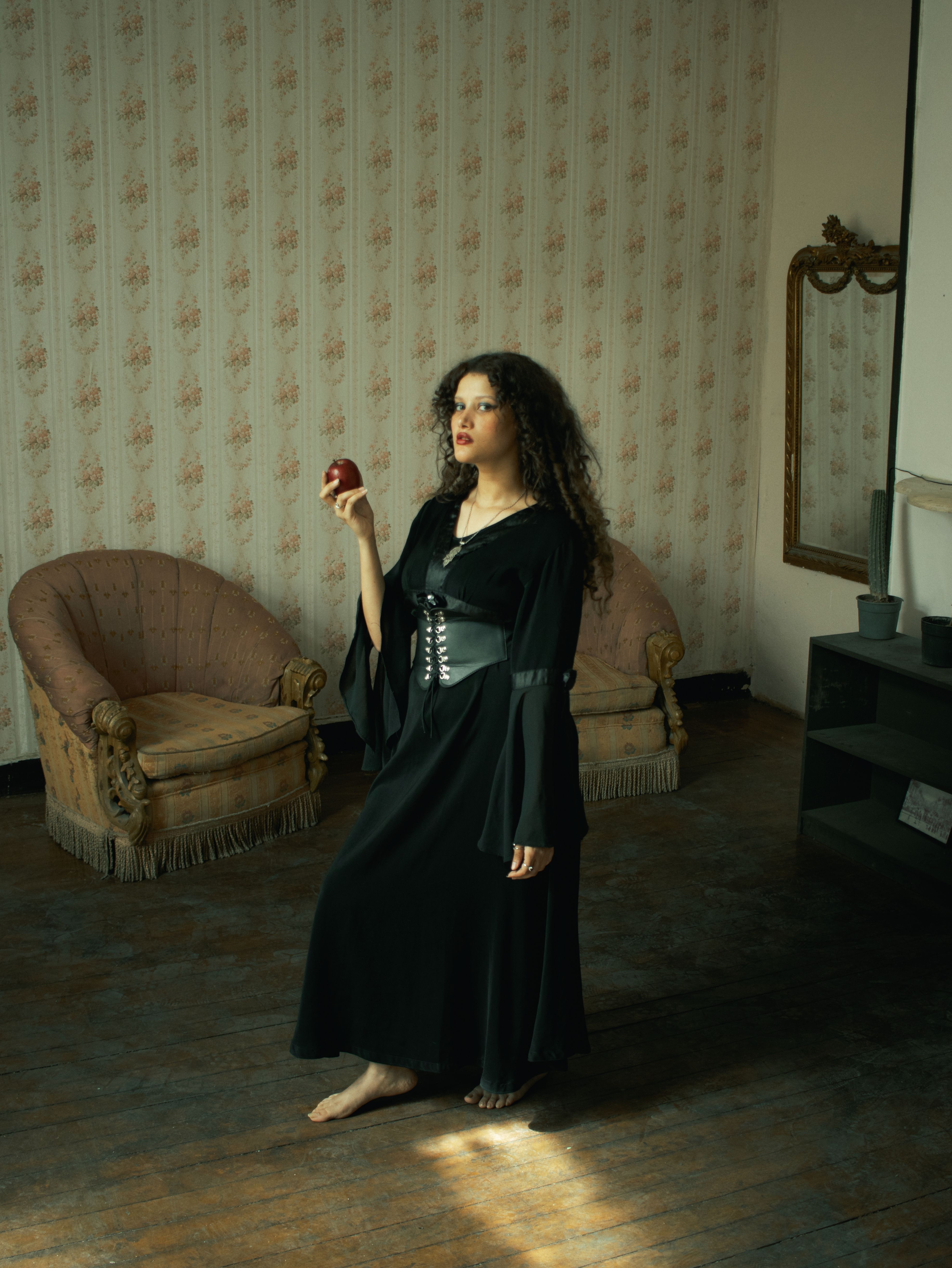 Woman in dark dress holding an apple in a vintage-inspired room. Unique and mysterious atmosphere.