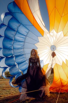 An elegant woman in a black dress poses near a vibrant blue and orange parachute outdoors.