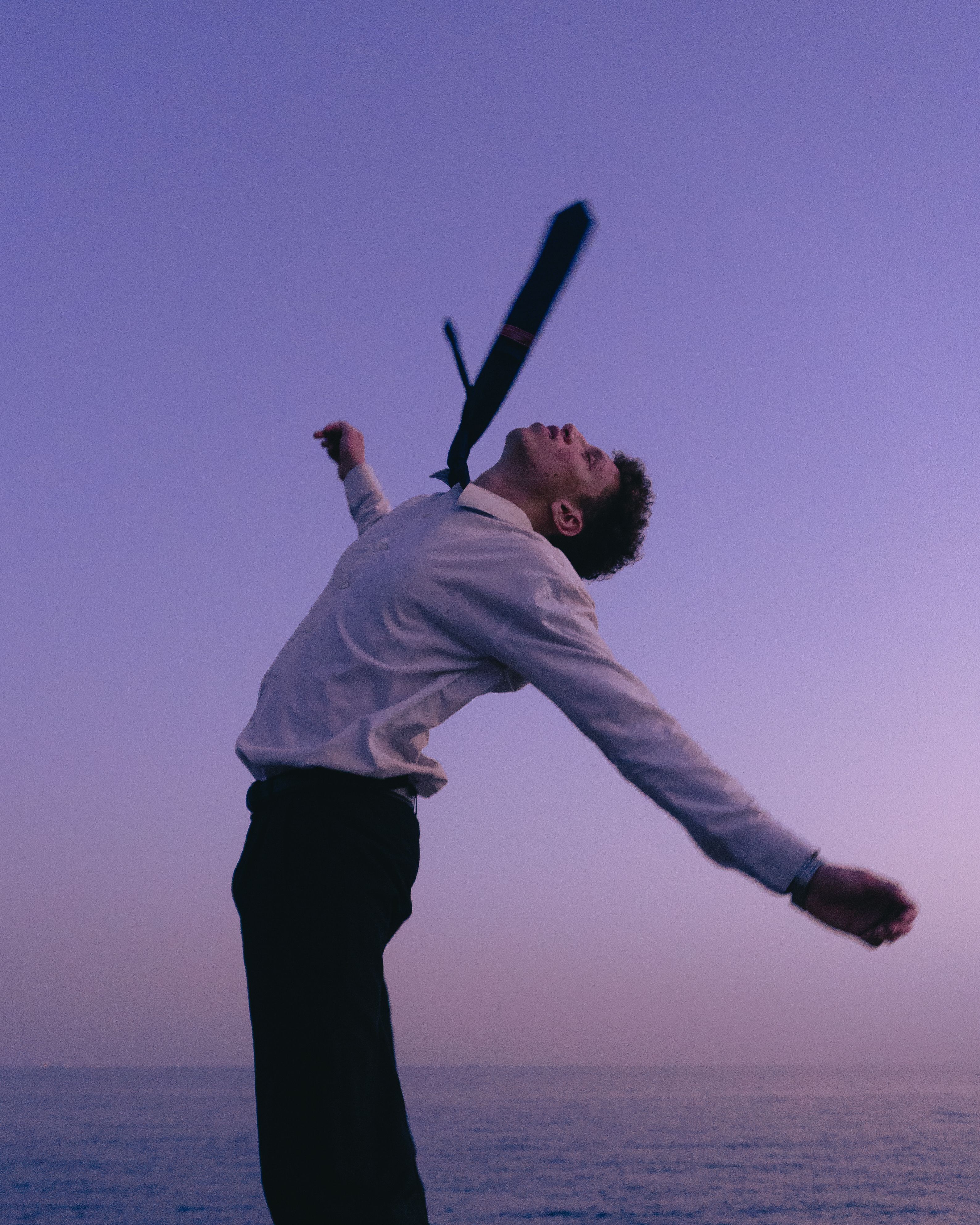 A young man joyfully throws his tie in the air by the sea at sunset, embodying freedom and happiness.