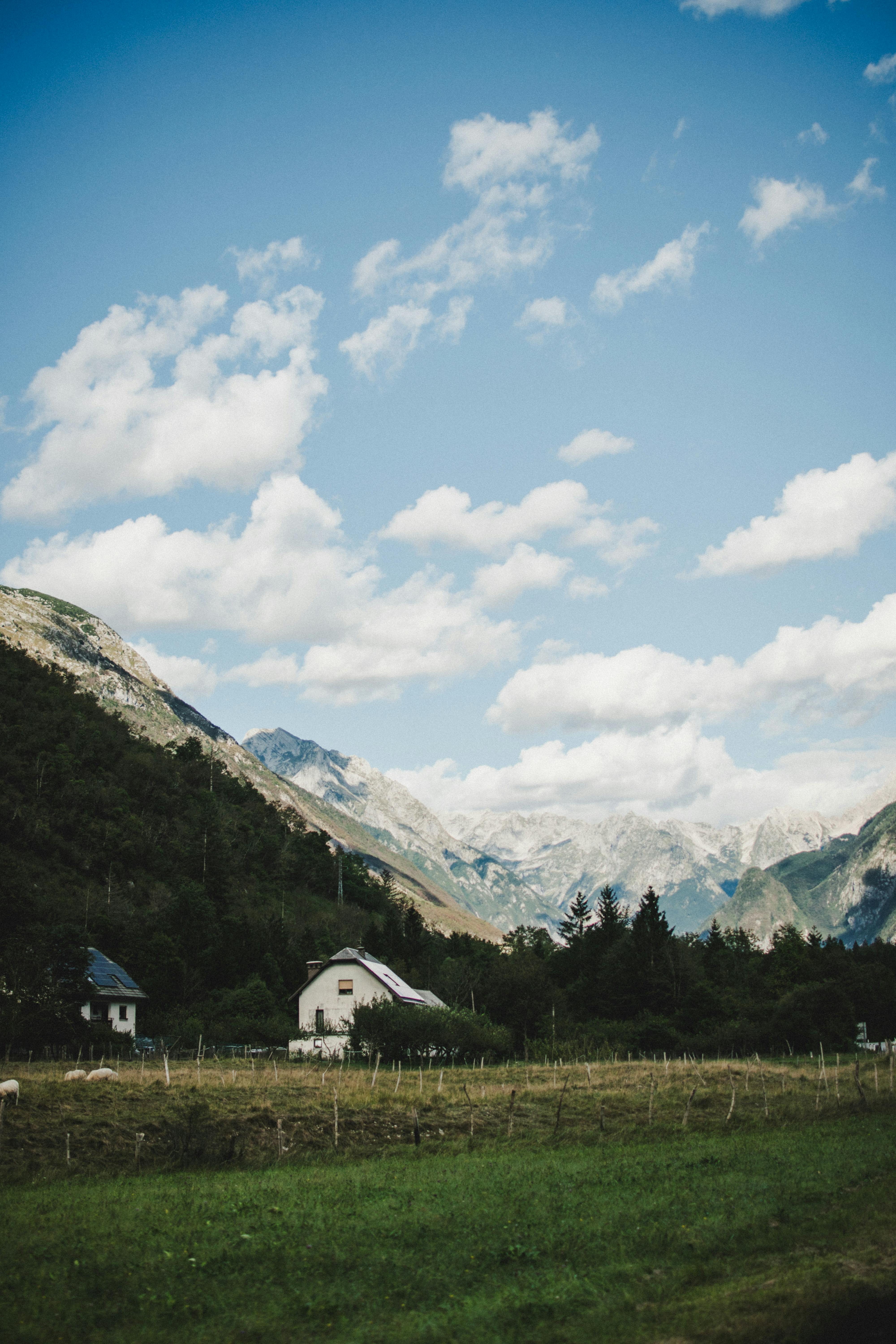 Picturesque farmhouse in a lush valley with dramatic mountains and a cloudy blue sky.
