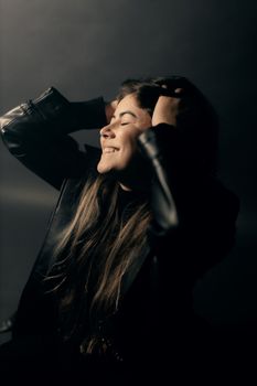 A woman in a leather jacket smiles contentedly with eyes closed, bathed in soft lighting.