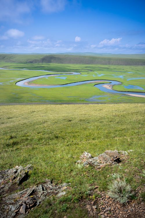Free A calm and scenic view of a winding river across lush green fields and blue sky. Stock Photo