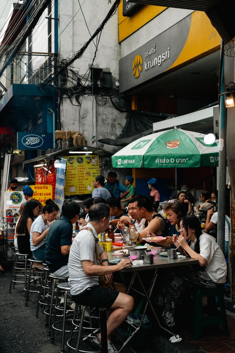 People eating at outdoor stools and tables on a busy Bangkok street lined with food stalls.