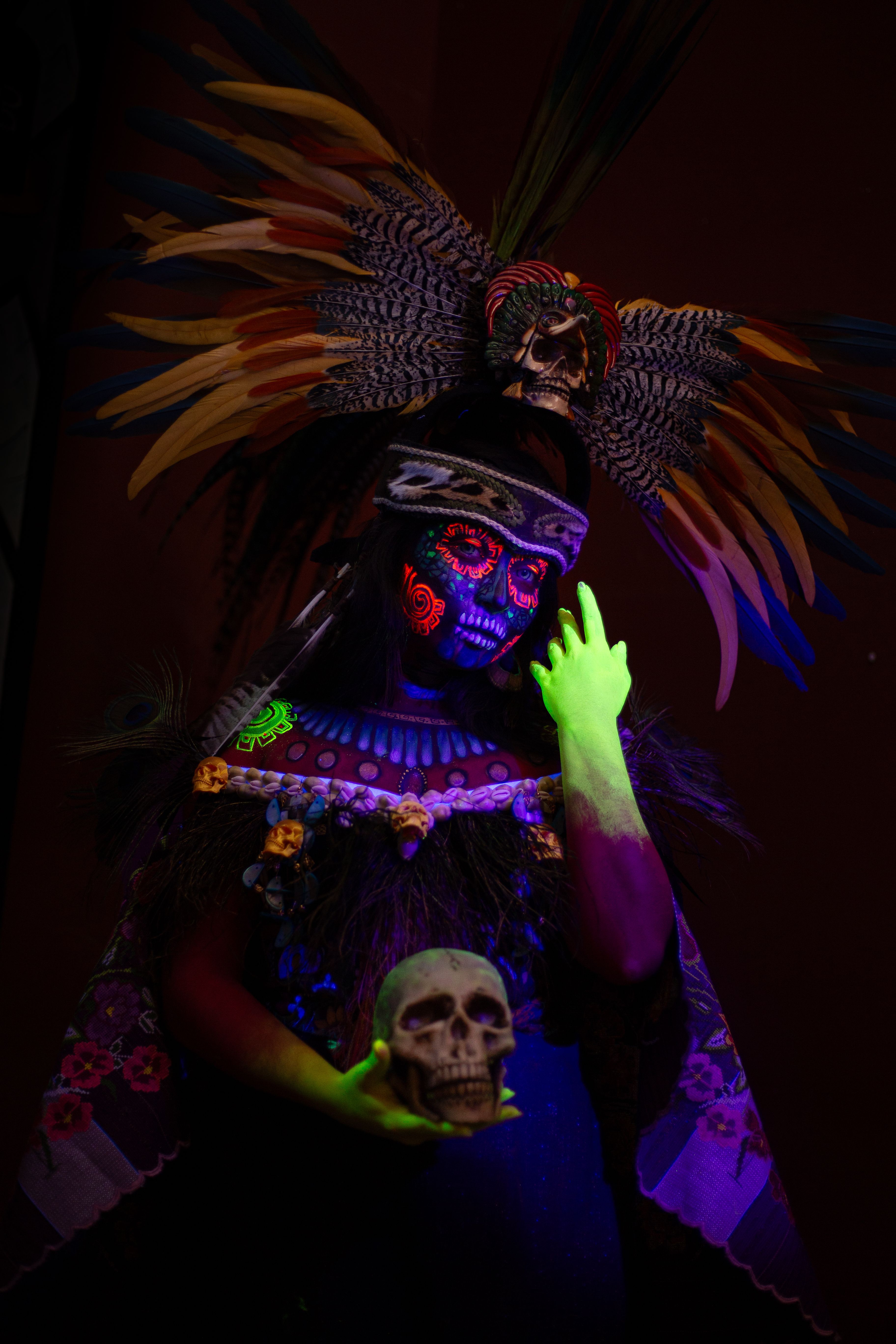 A striking portrayal of an Aztec-inspired Day of the Dead figure with fluorescent skull makeup and elaborate feathers.