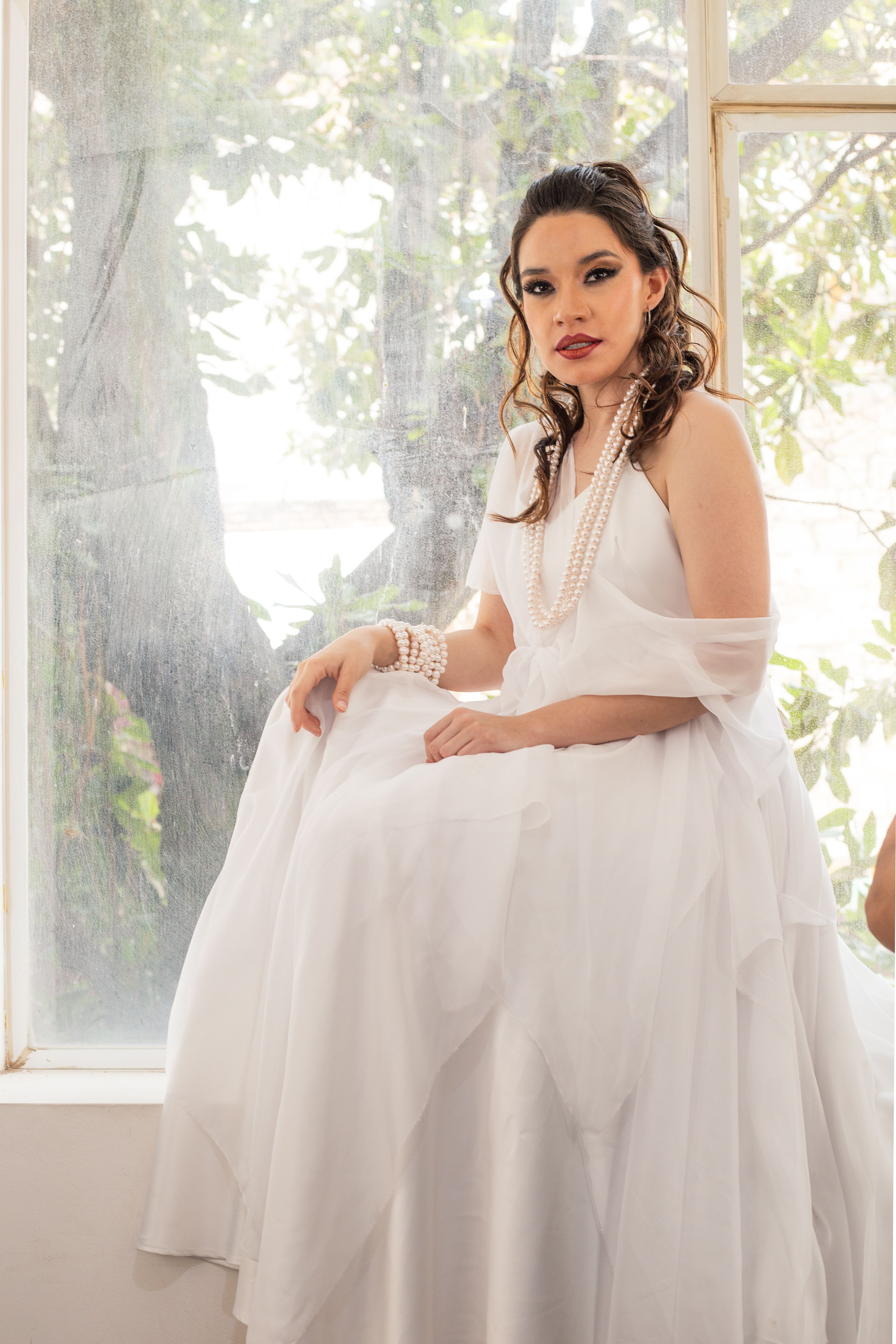 A beautiful woman in a white gown with pearls sitting by a window, bathed in sunlight.